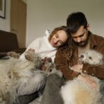A joyful couple hugging their two fluffy dogs while sitting indoors, showcasing love and companionship.